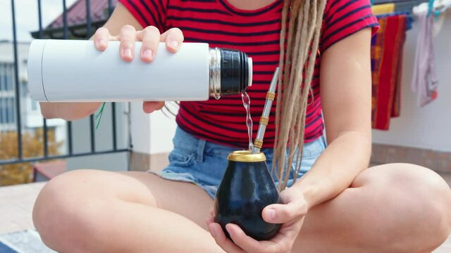 A woman brews Argentine mate tea. A traveler pours boiling water from a thermos into a calabash