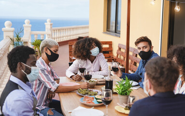 Obraz premium Cheerful young multiracial friends enjoy dinner at home on terrace while wearing protective face mask for coronavirus