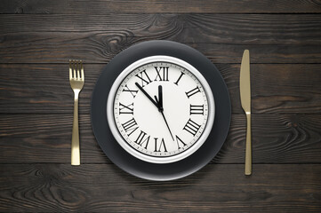 Festive table setting with black dish, clock, knife and fork on dark wooden table. New Year table setting. Flat lay, top view. Minimal composition