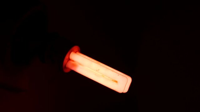 Fluorescent electric lamp glows faintly with pink light in dark room. End-of-life lamp with burnable electrodes. Previous generation energy saving lamp contain toxic mercury. Environment pollution