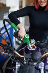 Obraz premium A slim woman fueling her modern motorcycle in refueling with gun. A refueling in sunny day. Custom urban bike.