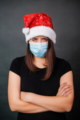 Portrait of a woman in a black T-shirt, a disposable medical mask and a Christmas hat on a gray background. Alone quarantine christmas celebration concept.