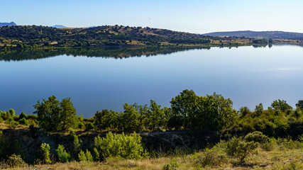 Vista aérea del lago con agua cristalina y reflejos de las montañas y del cielo azul, árboles en el primer plano.

