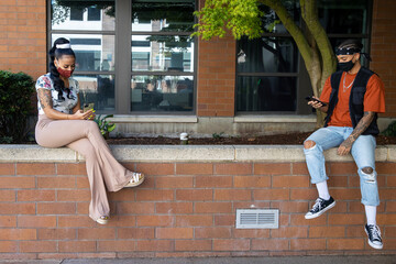 Two African American women wearing masks to protect from the Covid-19 coronavirus sit 6 feet apart for social distancing while looking at their smartphones outside