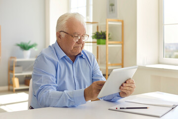 Senior man with glasses sitting at a table at home using a tablet reads information on the Internet.