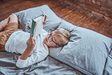 Dressed in casual clothing cheerful and smiley boy reads book lying down on a bed in living room. © Fxquadro