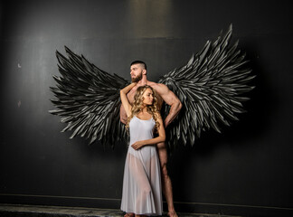 handsome muscular man in angel costume with black wings and fragile defenseless beautiful girl in...