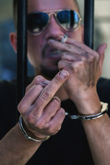 Upset handcuffed man with cigarette imprisoned for crime, punished for serious villainy. He showing middle finger gesture. Arrest, gangster, deviant behavior concept.