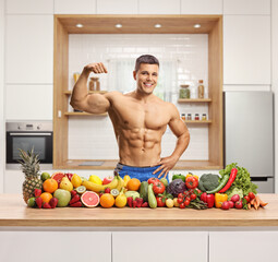 Naklejka premium Muscular man flexing biceps muscle and posing with a pile of fruits and vegetables on a kitchen counter