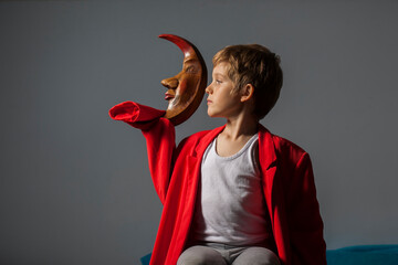 niño rubio con abrigo rojo sujeta luna de madera roja con la mano mirando de perfíl
