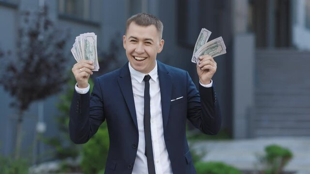 Man shows money and celebrating success, victory while looking to camera. Outdoors. Amazed happy excited businessman with money - U.S. currency dollars banknotes