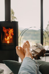Feet at modern fireplace and window with view on mountains. Woman barefoot relaxing in cozy home