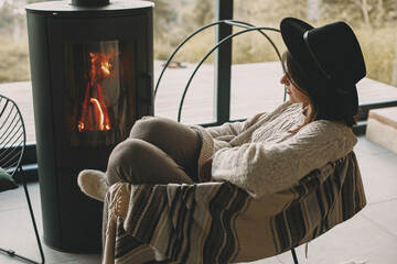 Stylish woman in knitted sweater and hat warming up at modern black fireplace. Cozy home