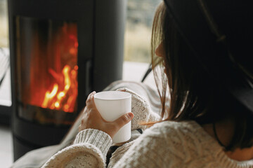 Stylish woman in knitted sweater relaxing with warm cup of tea at modern fireplace, closeup