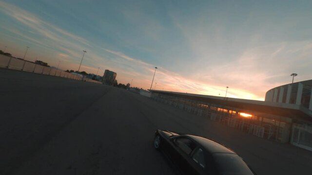 Black jdm car driving on urban on golden sunset landscape, aerial view. Fpv cinematic drone shooting modern automobile riding on twilight street on evening sunlight through city buildings.