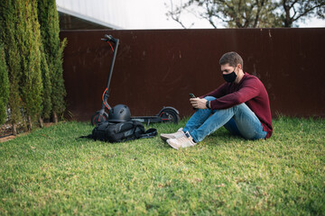 Fototapeta premium Young man using a mobile phone with an electric skate sitting on the grass.