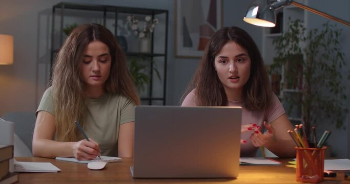 Portrait of two Caucasian twin sisters sitting in room and studying while having online lesson on laptop Pretty female writing in notebook while sister talking on video on computer distance education