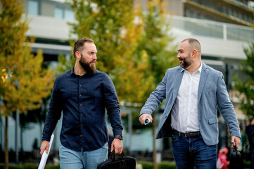 Satisfied business colleagues discussing project on their way to office. Business men walking outside, holding papers, talking, laughing. 