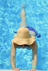 a girl in a wide-brimmed straw beach hat holds her hands over the side of the pool. blue water. in a swimsuit.