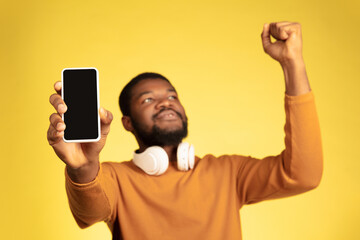 Showing phone screen, winner. Young african-american man's portrait on yellow studio background, facial expression. Beautiful male model with headphones, copyspace. Concept of human emotions, sales