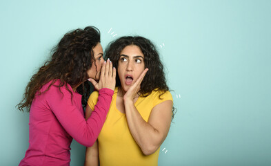 Woman speaks in the ear to a friend about a great promotion. amazed expression. cyan background
