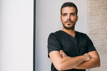 Male dentist in black medical scrubs looking away while standing with arms crossed against wall