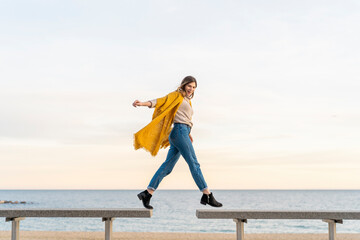 Cheerful young woman hopping from bench to another at beach promenade against sky during sunset