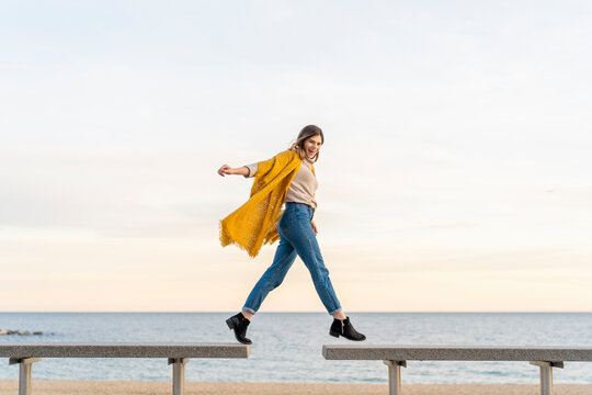 Cheerful young woman hopping from bench to another at beach promenade against sky during sunset