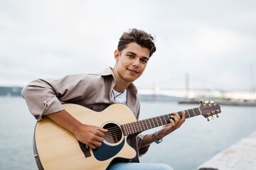 Handsome young man playing guitar while practicing at promenade against sky