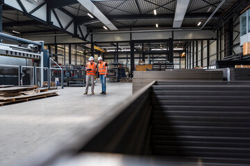 Two men wearing hard hats and safety vests talking on factory shop floor