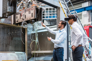 Man explaining machine to colleague wearing VR glasses in factory