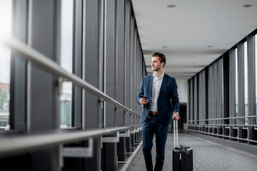 Young businessman in a passageway with cell phone, earbuds and rolling suitcase on the go