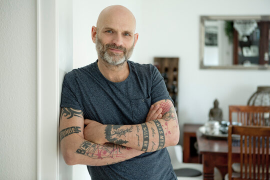 Tattooed man leaning in door of his home, with arms crossed
