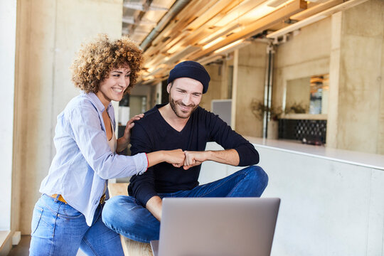 Happy man and woman with laptop fist bumping in modern office