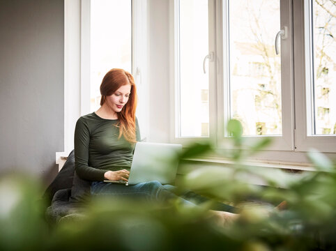 Redheaded woman sitting beside  window using laptop