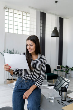 Smiling young female architect holding plan while sitting at desk in creative office