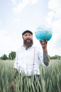 Bearded man holding globe looking away while standing amidst cornfield against sky