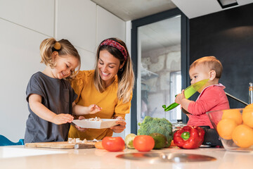 Smiling mother and girl preparing food while baby daughter sitting on kitchen island