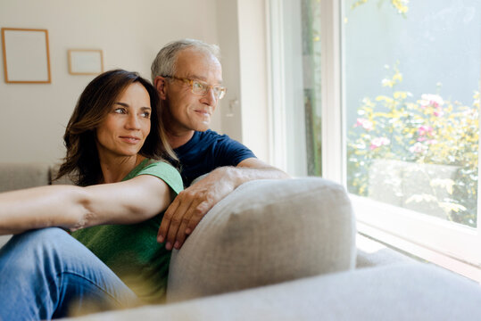 Smiling Mature Couple Sitting On Couch At Home Looking Out Of Window