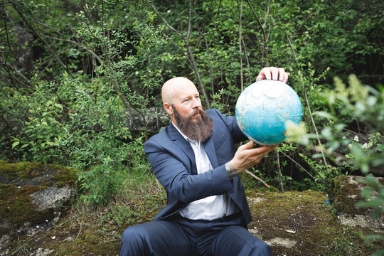 Bald bearded businessman holding globe while sitting against trees in forest