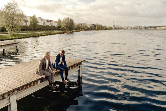Two businessmen sitting on jetty at a lake talking