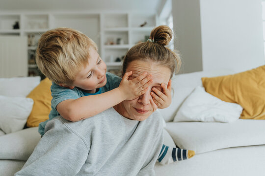 Boy covering eyes of mother from behind while sitting by sofa at home