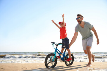 Happy father teaching son to ride bicycle on sandy beach near sea