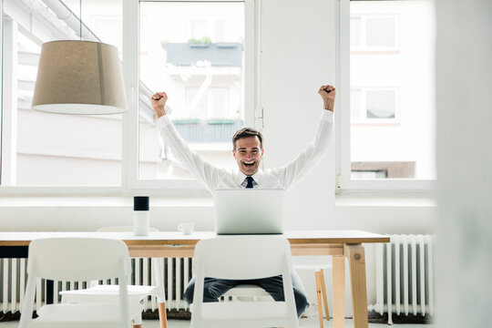 Laughing businessman with laptop on table cheering in office