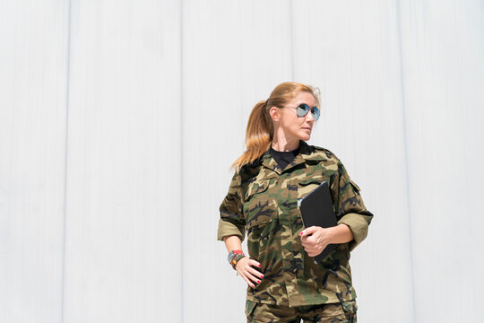 Mature blond female army soldier wearing sunglasses while holding digital tablet and looking away at military base on sunny day
