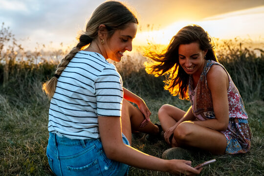 Smiling young woman showing smart phone to female friend while sitting on field during sunset