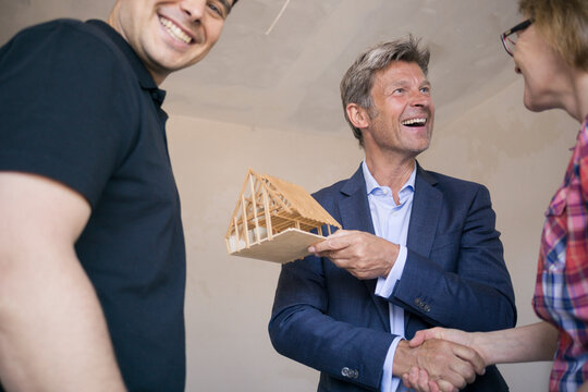 Happy man in suit holding model house shaking hands with couple