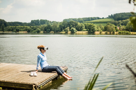 Woman with VR glasses sitting on jetty at a lake with feet in water