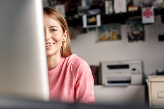 Portrait of smiling young woman behind computer screen at desk at home