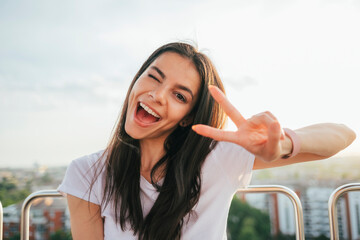 Cheerful beautiful woman showing peace sign while enjoying Ferris wheel against sky at sunset